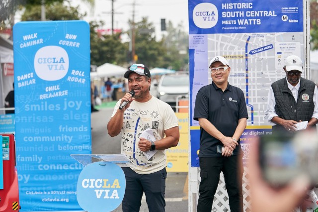Man in tan shirt and baseball hat speaking into a microphone at a press conference for a city-wide bicycling event.